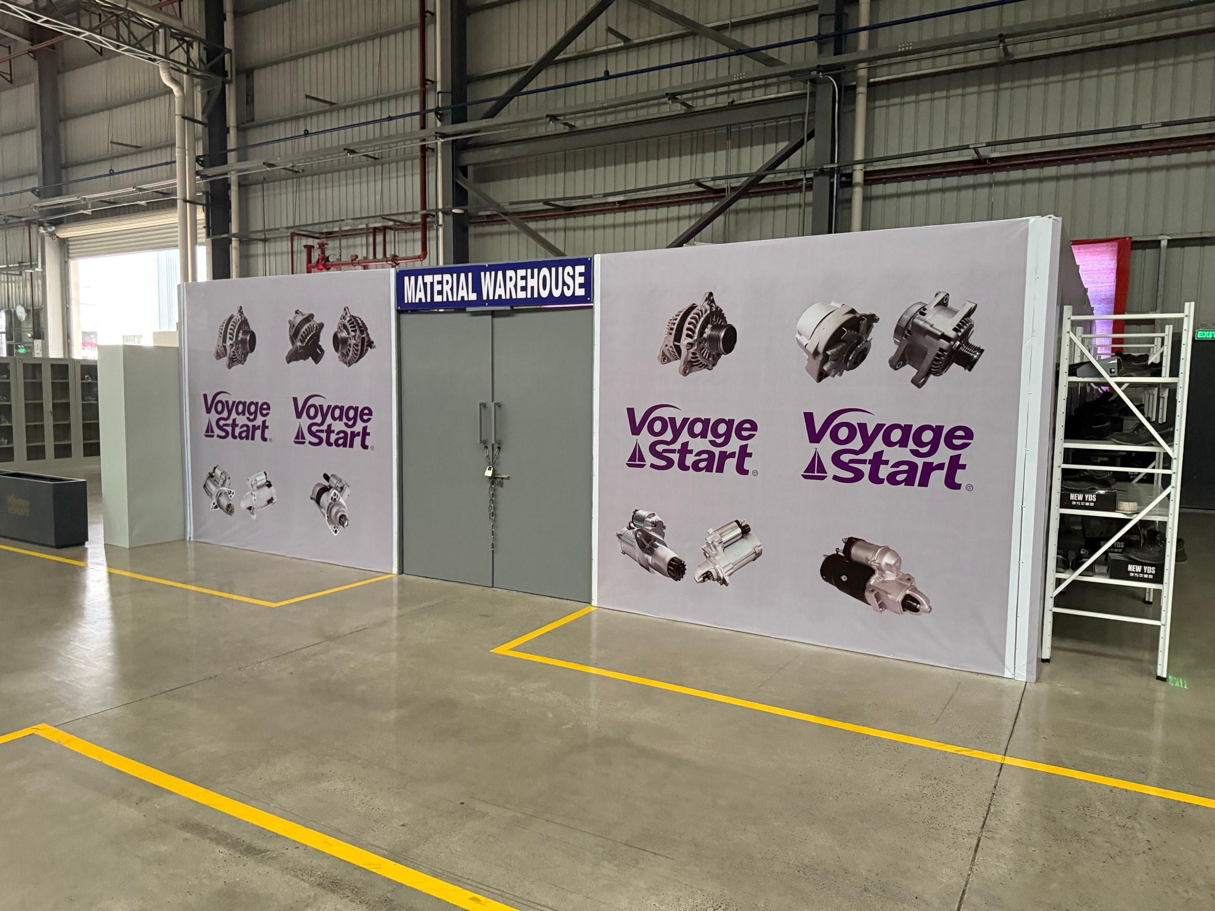 The material warehouse inside the plant: a walled stock room with a locked double door under a Material Warehouse sign, VoyageStart banners on the walls and a rack of boxed parts beside it.