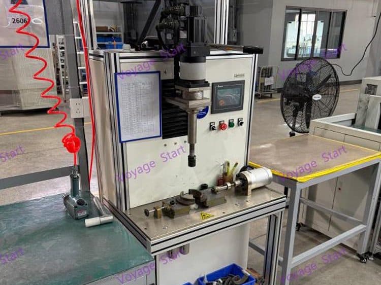 A vertical press with a touchscreen control panel above a tooling fixture, with an alternator on the table beside it.