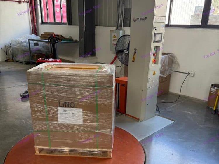 A stretch-wrapped pallet of cartons, strapped and labeled, on the turntable of a pallet wrapping machine.